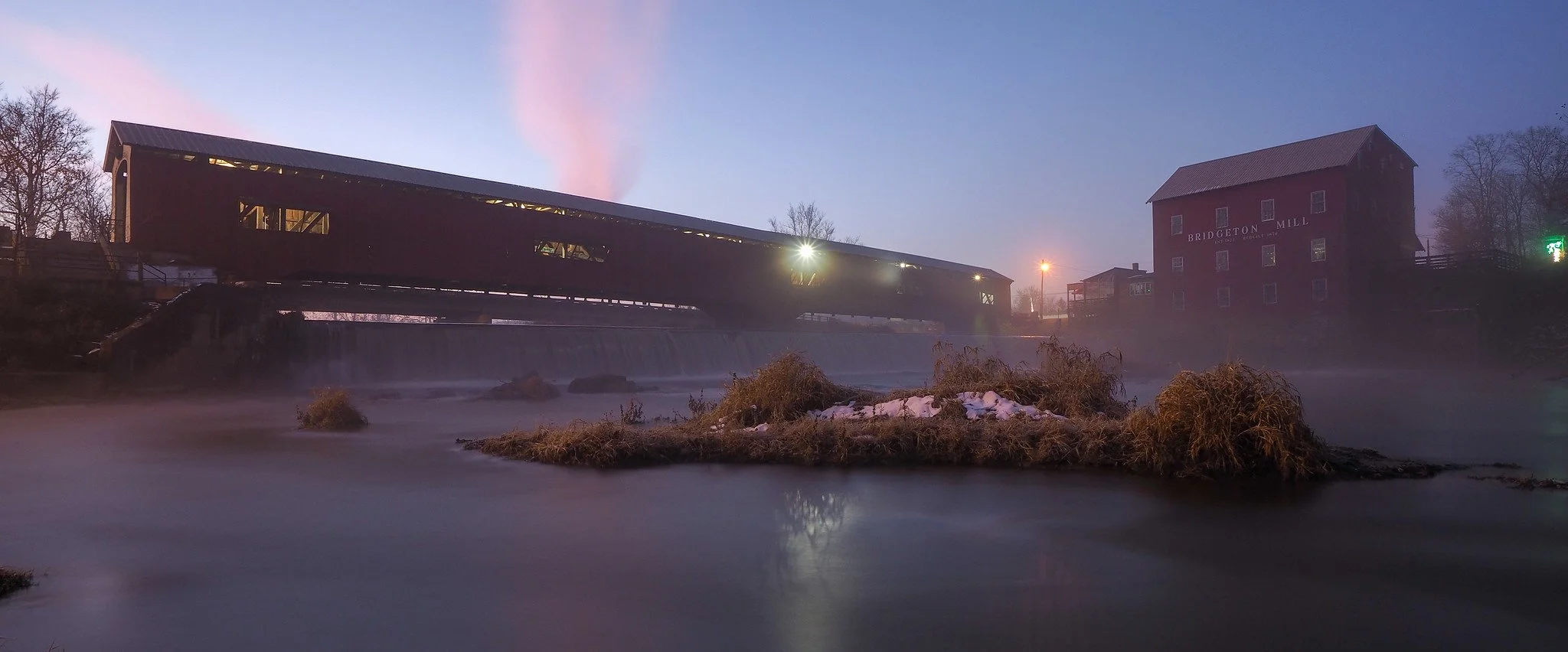 A covered bridge over a river at dusk with a traditional mill building on the right side, labeled 'Bridgeton Mill.' The scene includes calm water, some rocks and snow, and sparse trees in the background.