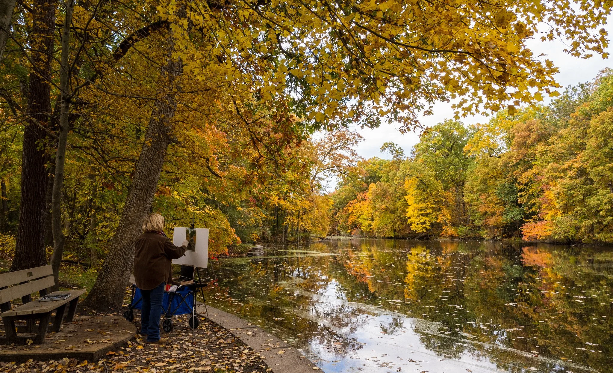 A woman painting outdoors along a river during fall, with trees displaying orange, yellow, and green leaves, and their reflection on the water.