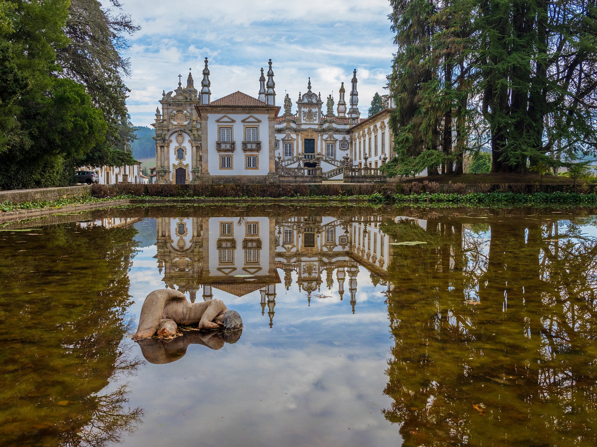 A historic ornate white and brown palace with multiple spires, seen through a reflecting pond surrounded by trees. The sky is partly cloudy.