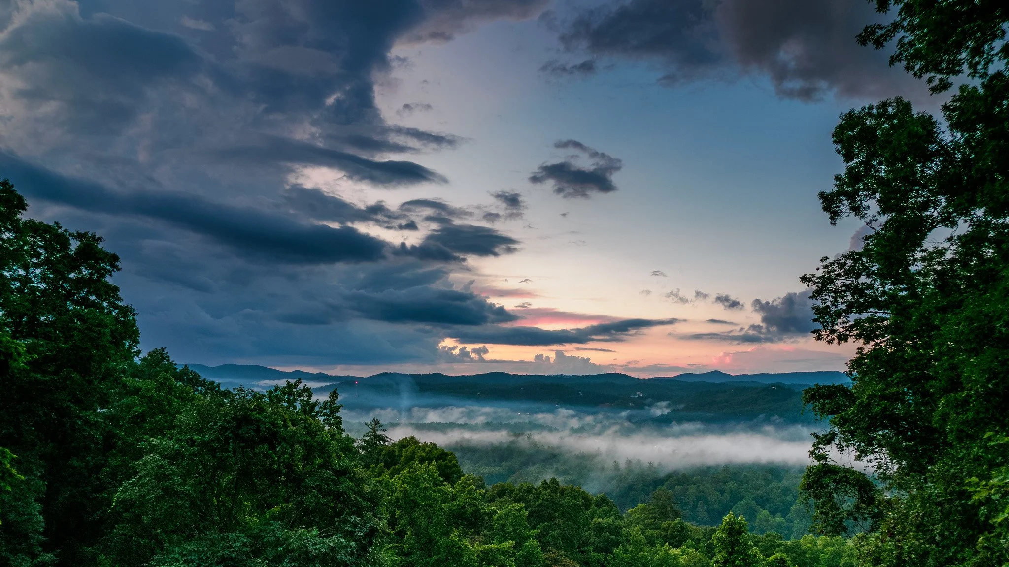 Dramatic sky over a lush green forest with mist in the valleys and mountains in the distance at sunset or sunrise.