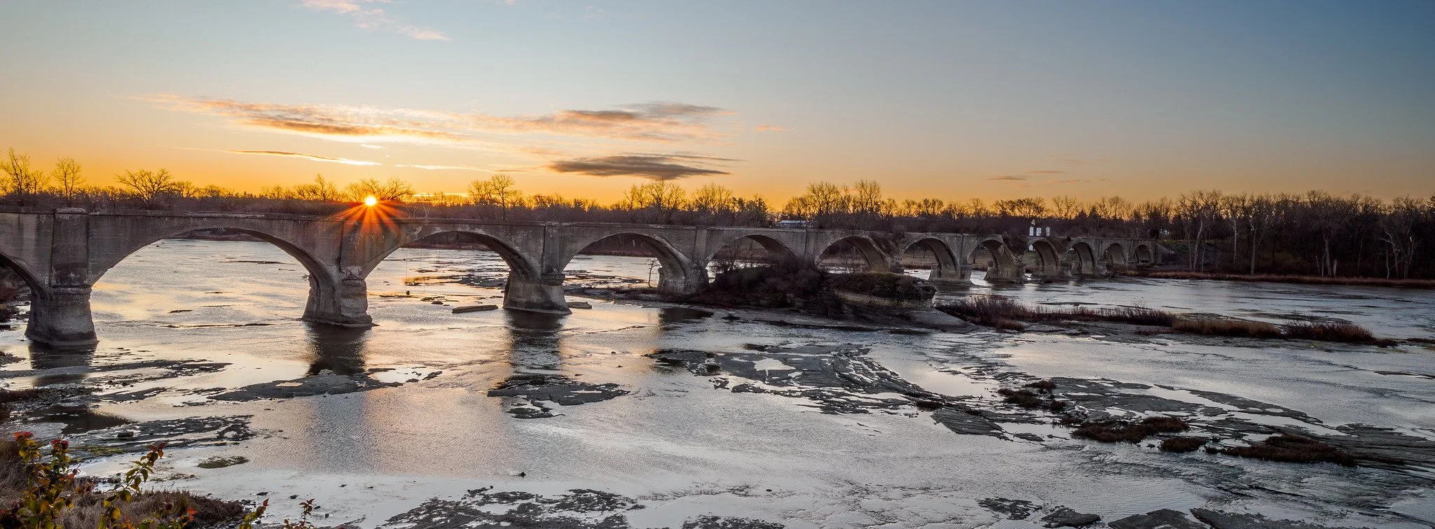 Sunset over a stone bridge crossing a partially frozen river, with leafless trees along the banks and a clear sky with some clouds.