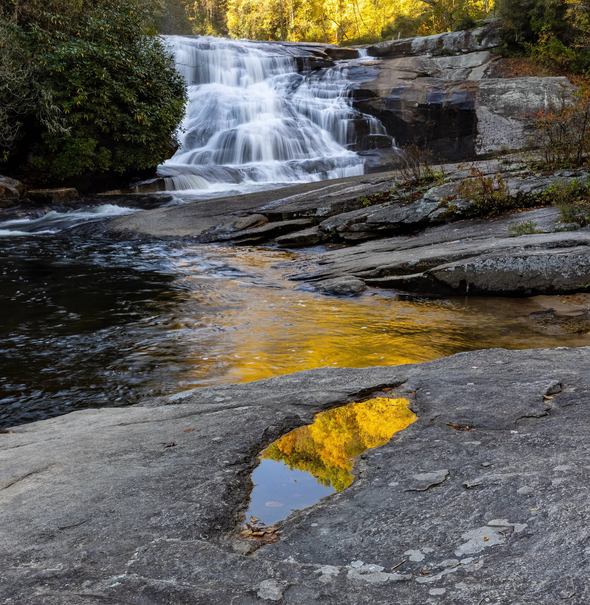 A scenic view of a waterfall flowing over rocks, with a small pool of water reflecting autumn trees and sky in the foreground.