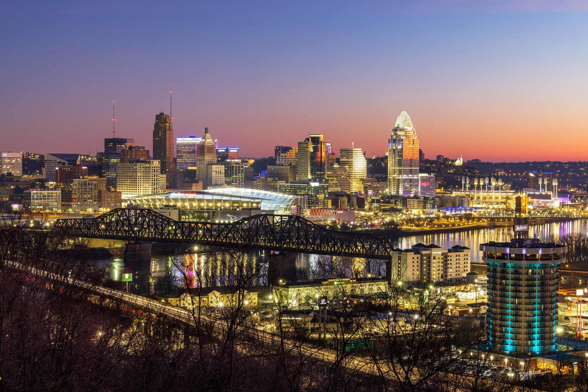 Nighttime cityscape of Cincinnati, Ohio, featuring illuminated skyscrapers, the Ohio River, and a bridge, with a colorful sunset sky in the background.