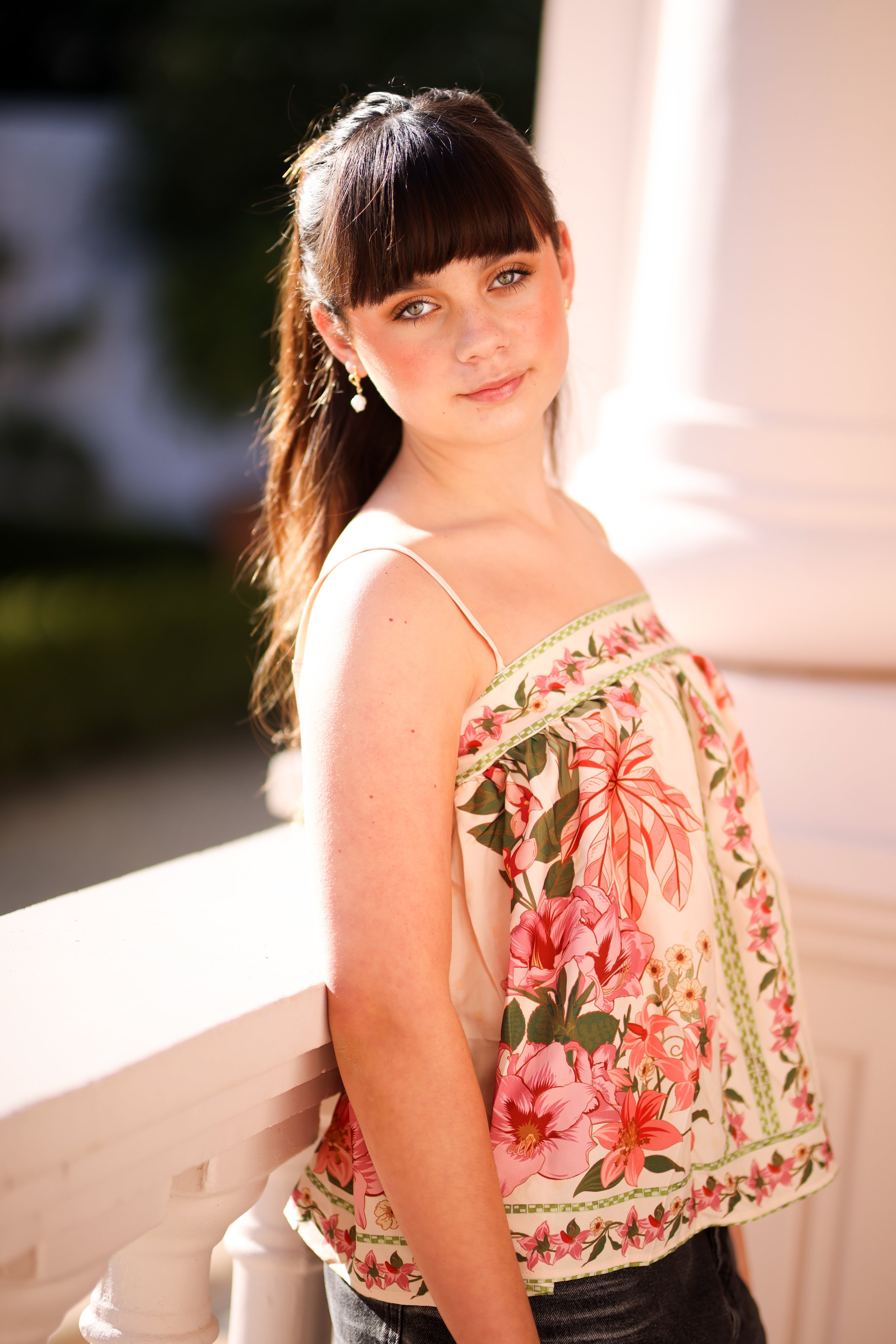 A young girl with brown hair and bangs posing outdoors in sunlight, wearing a floral sleeveless top and earrings.