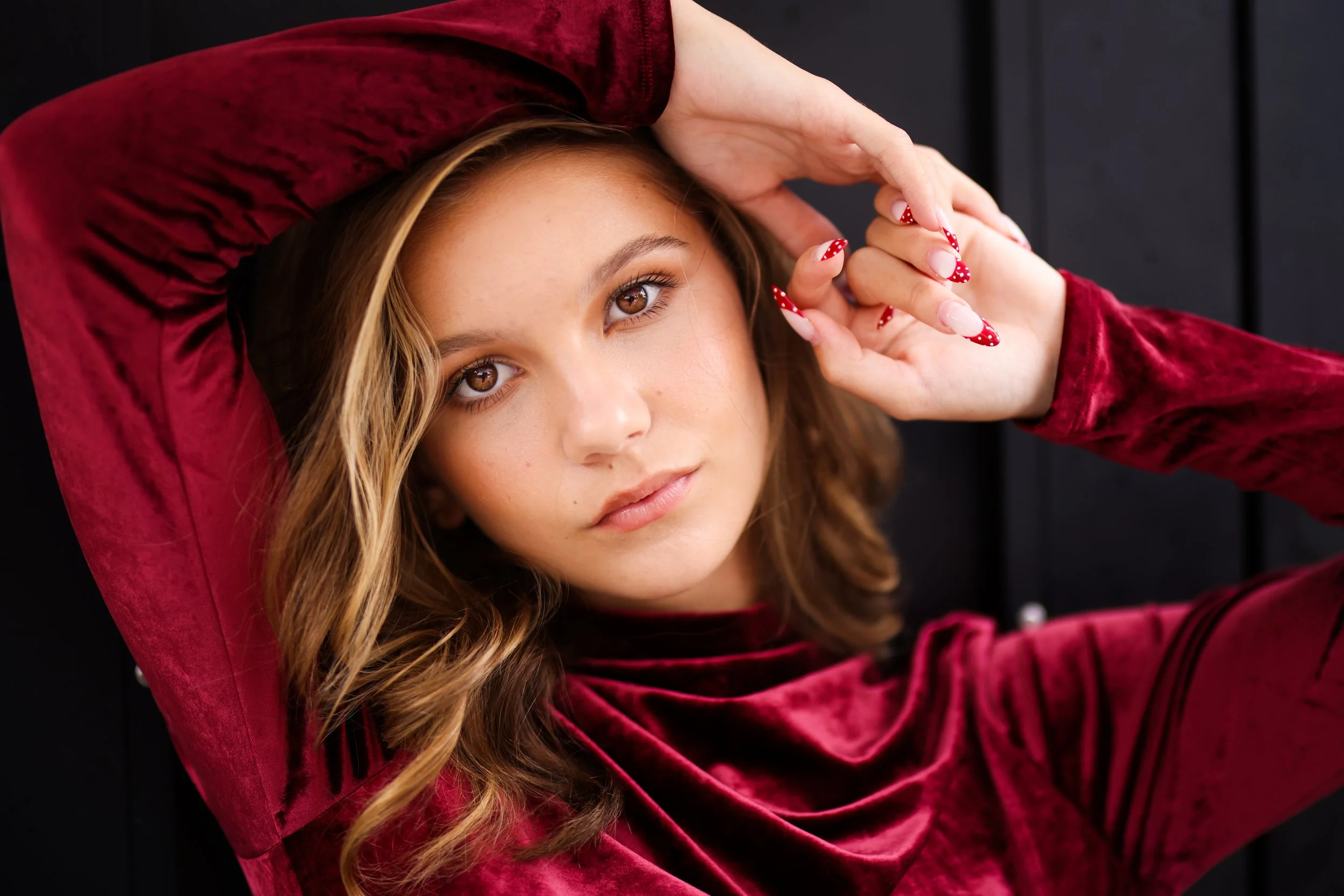 A young woman with wavy brown hair wearing a red velvet long-sleeve top, with her hand resting on her head, looking directly at the camera against a dark background.