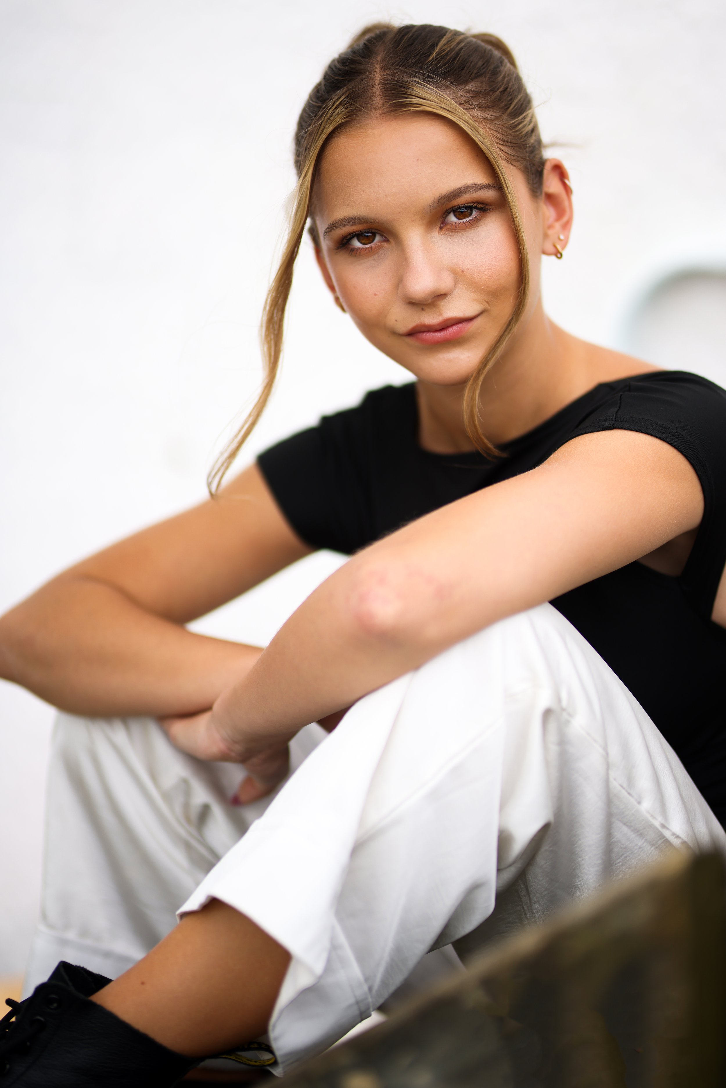 A young woman with blonde hair in a braid, wearing a black t-shirt and white pants, sitting with her arms resting on her knees against a plain white background.
