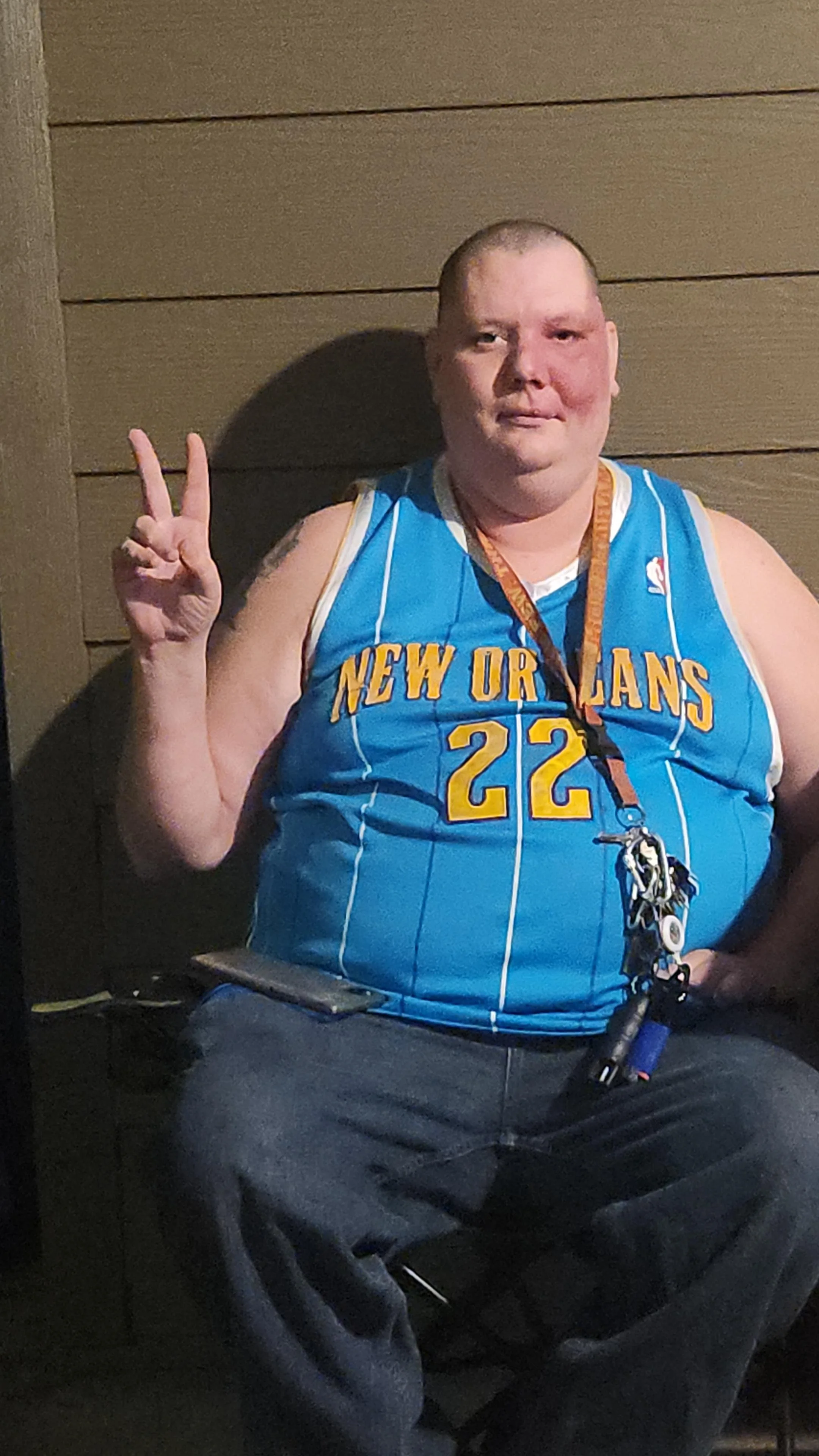A man with short hair and tattoos, wearing a blue New Orleans basketball jersey with the number 22, sits against a wooden wall, making a peace sign with his right hand and holding a smartphone in his lap.