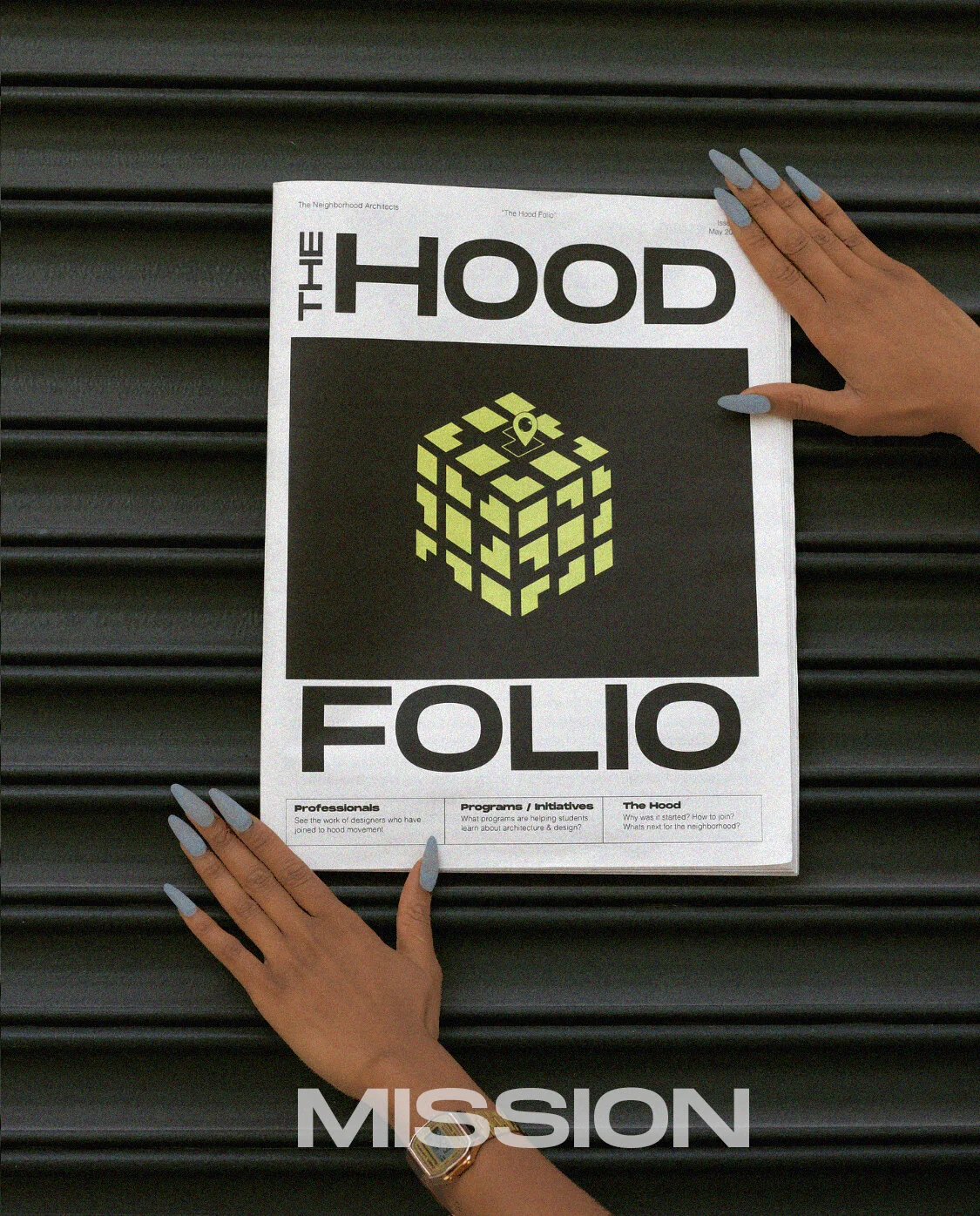 A person is holding a magazine titled "The Hood Folio" against a black grooved background. The cover features a stylized cube with a location pin icon and the words "professionals," "programs / initiatives," and "The Hood". The word "MISSION" is prin