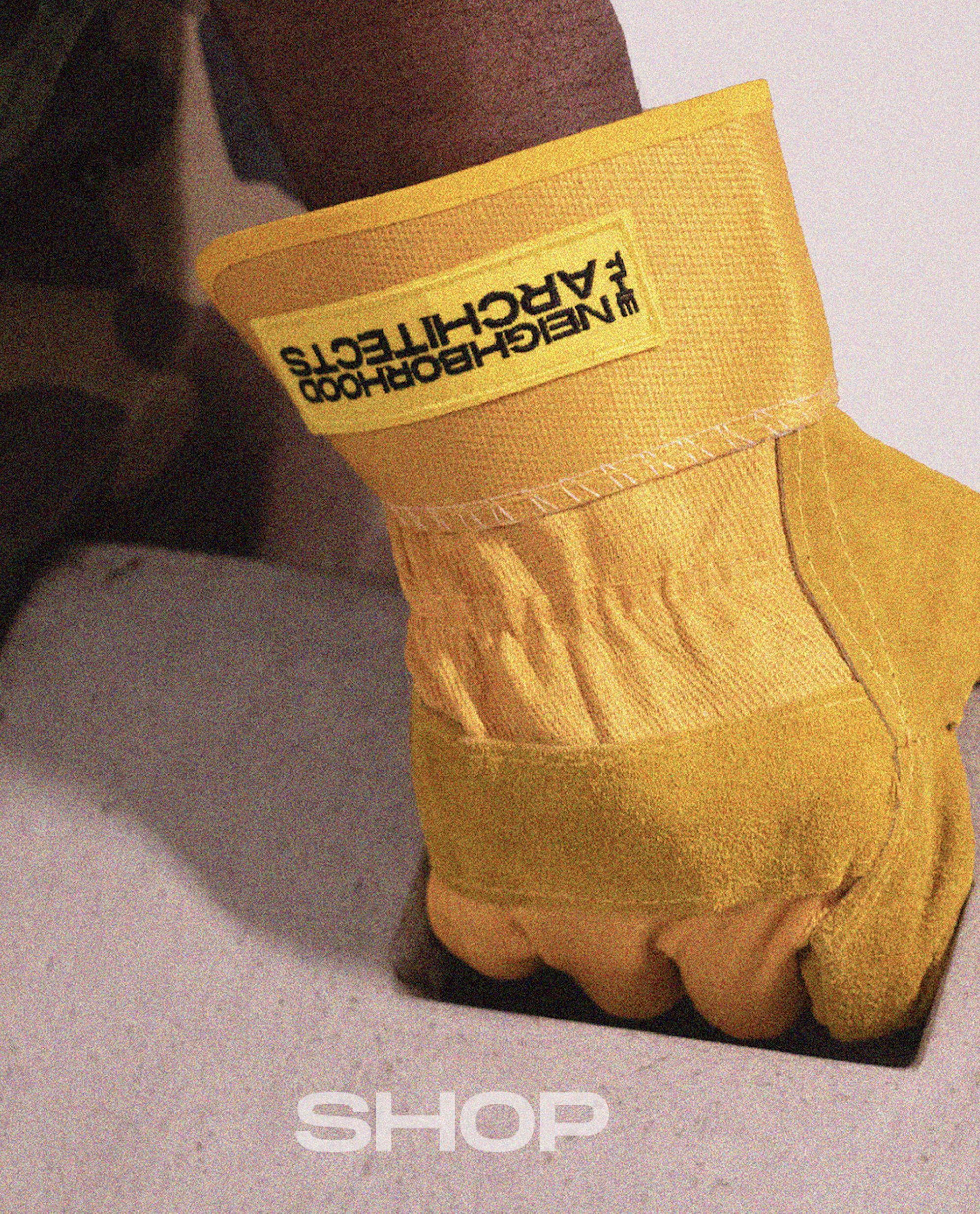 A close-up of a person's hand wearing a yellow glove with the text "Neighborhood Architects" on the cuff, resting on a concrete surface. The word "SHOP" is visible at the bottom of the image.