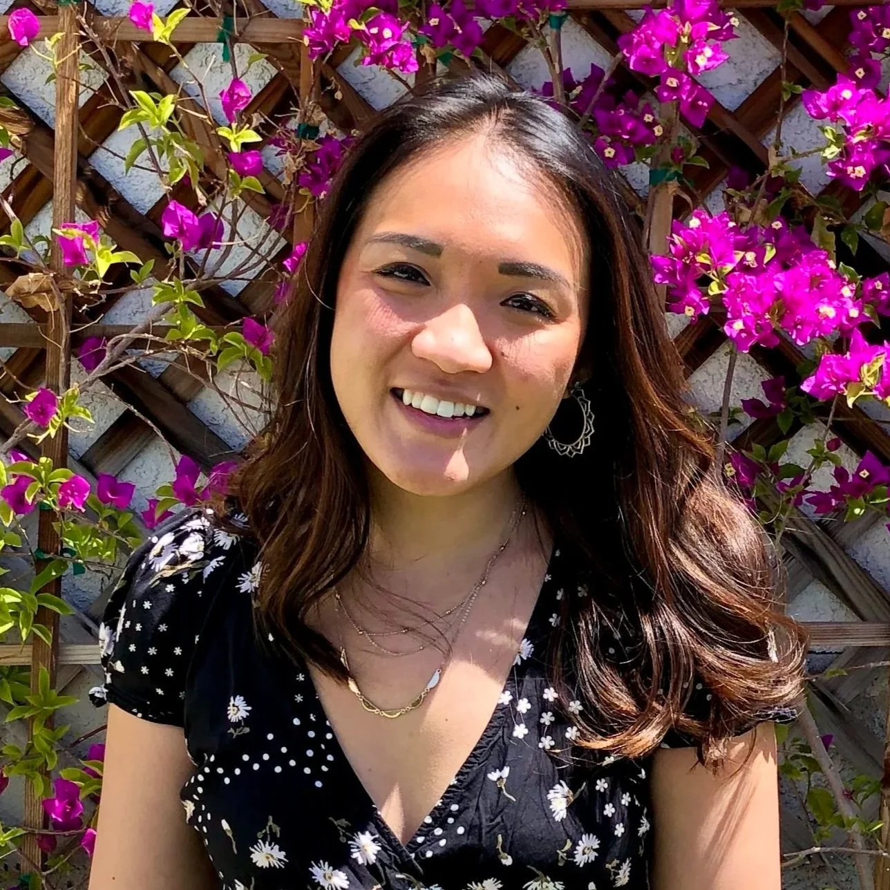 A smiling person with long brown hair, wearing hoop earrings, layered necklaces, and a black dress, standing in front of a lattice fence with vibrant pink flowers and green leaves.