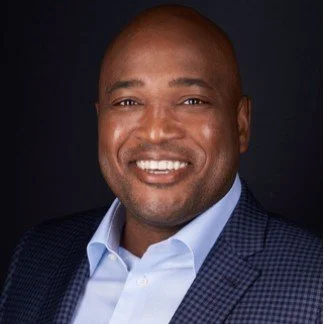 A smiling Black man in a blue suit and light blue shirt against a dark background.