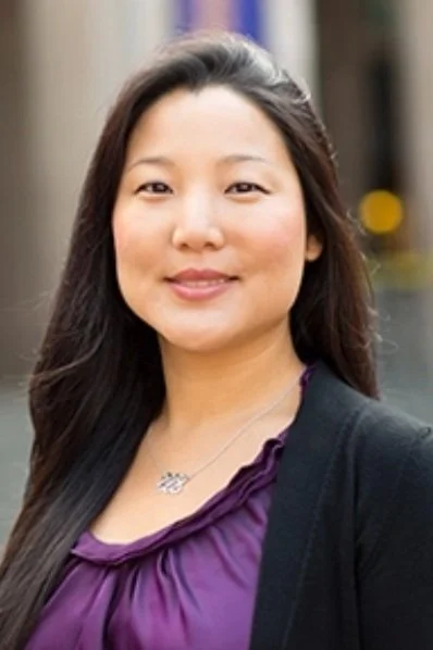 A woman with long dark hair wearing a purple blouse and black blazer, smiling outdoors.