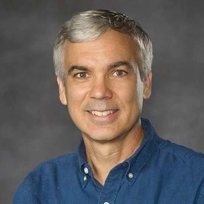 Portrait of a middle-aged man with gray hair, wearing a blue button-up shirt, smiling at the camera.
