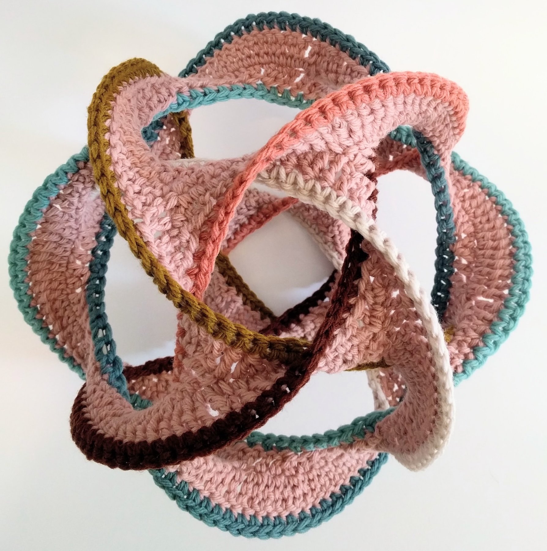 A colorful, intertwined crochet sculpture made of pink, brown, beige, teal, and coral yarns forming a complex 3D geometric shape.