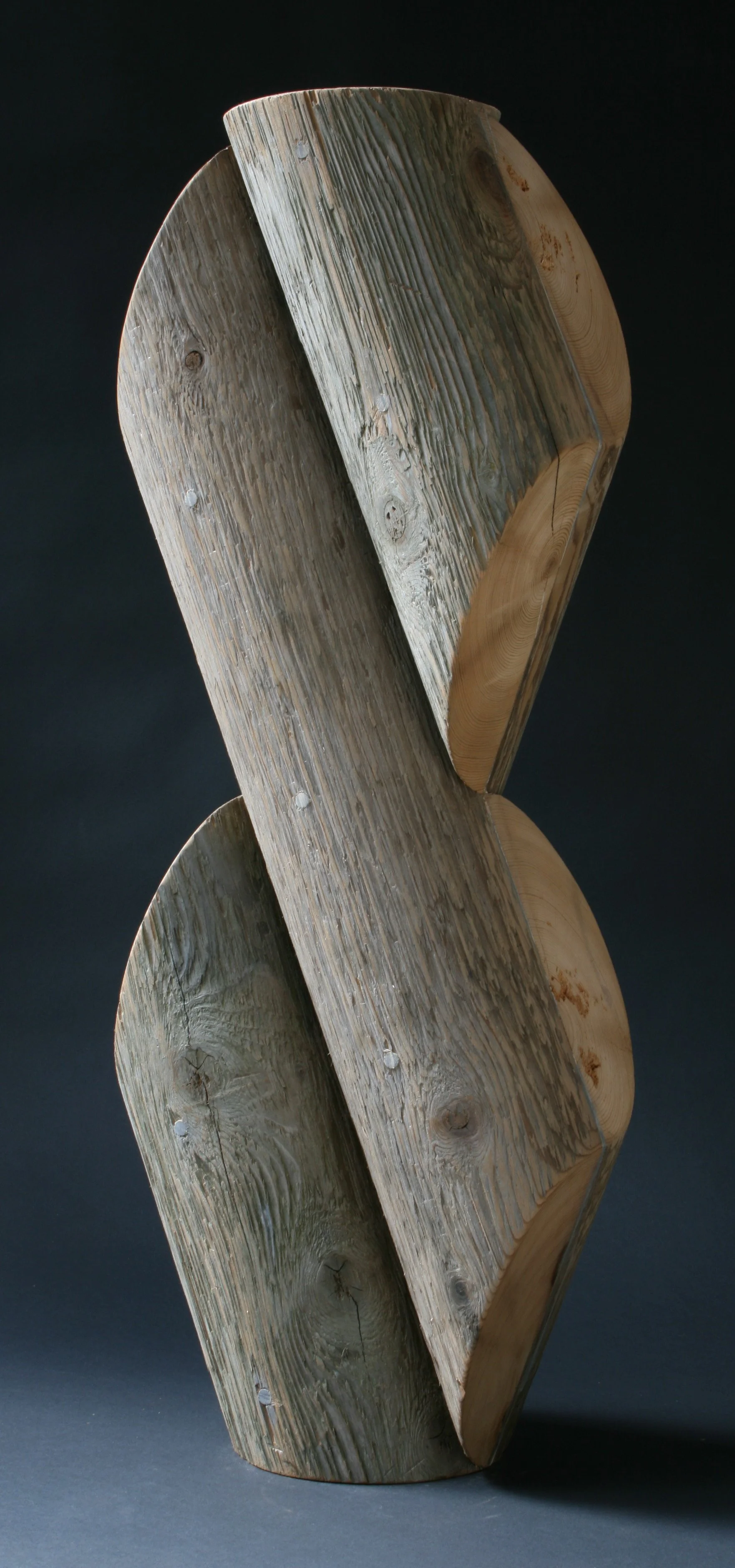 A modern abstract sculpture made of weathered wood with curved, overlapping shapes against a dark background.