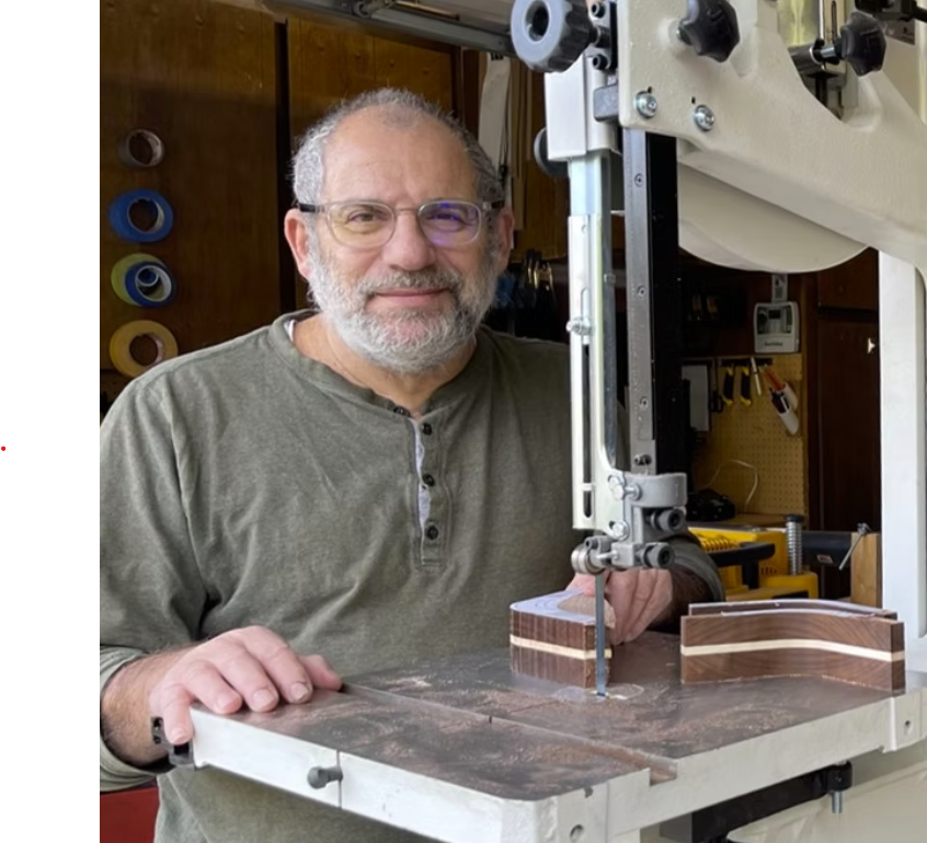 A man with glasses and a beard operating a scroll saw in a woodworking workshop. Wood pieces with layered color patterns are on the table.