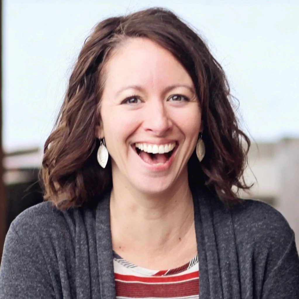 A woman with shoulder-length curly brown hair smiling widely, wearing earrings and a dark sweater over a striped shirt.