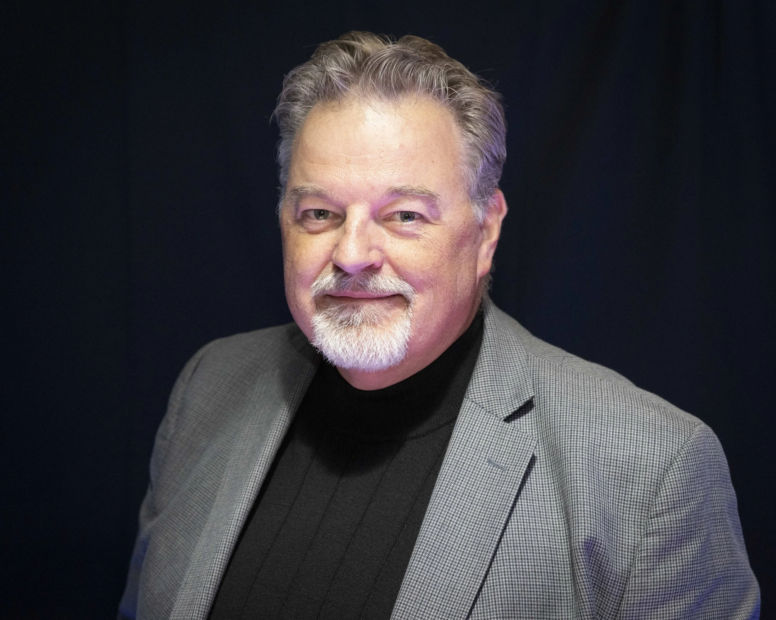 A smiling middle-aged man with gray hair and a beard, wearing a gray blazer and black turtleneck, against a dark background.