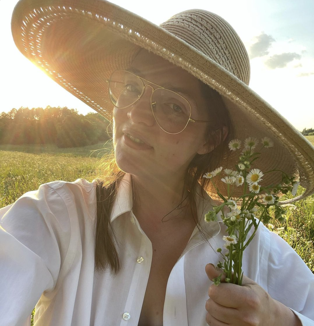 Woman wearing a large sunhat, glasses, and a white shirt, holding a small bouquet of daisies, standing in a sunny field at sunset.