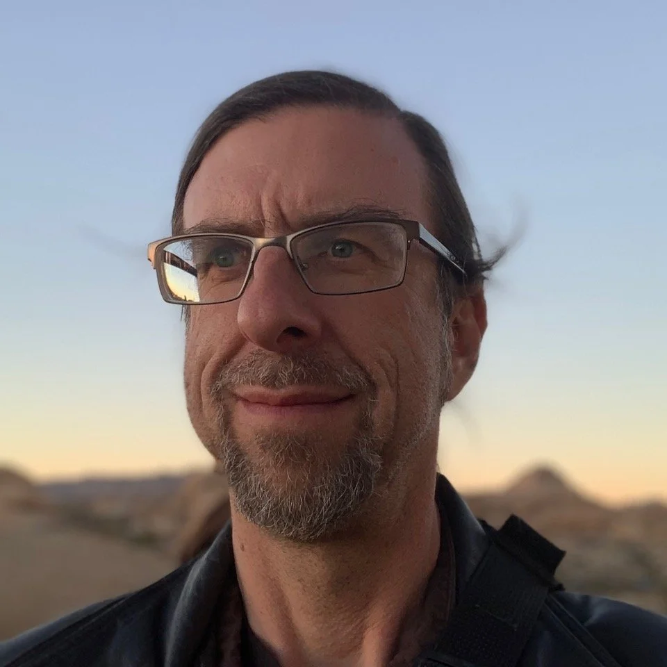 A man with glasses, a goatee, and shoulder-length hair outdoors during sunset or sunrise, with a blurred desert landscape in the background.