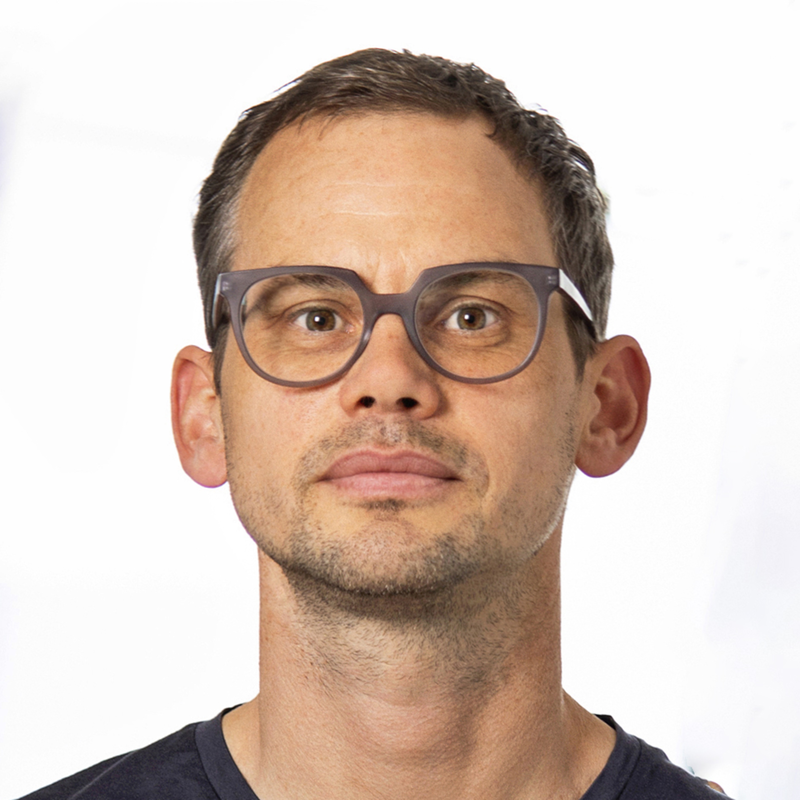 A man wearing glasses with brown hair, light facial stubble, and a serious expression, against a plain white background.