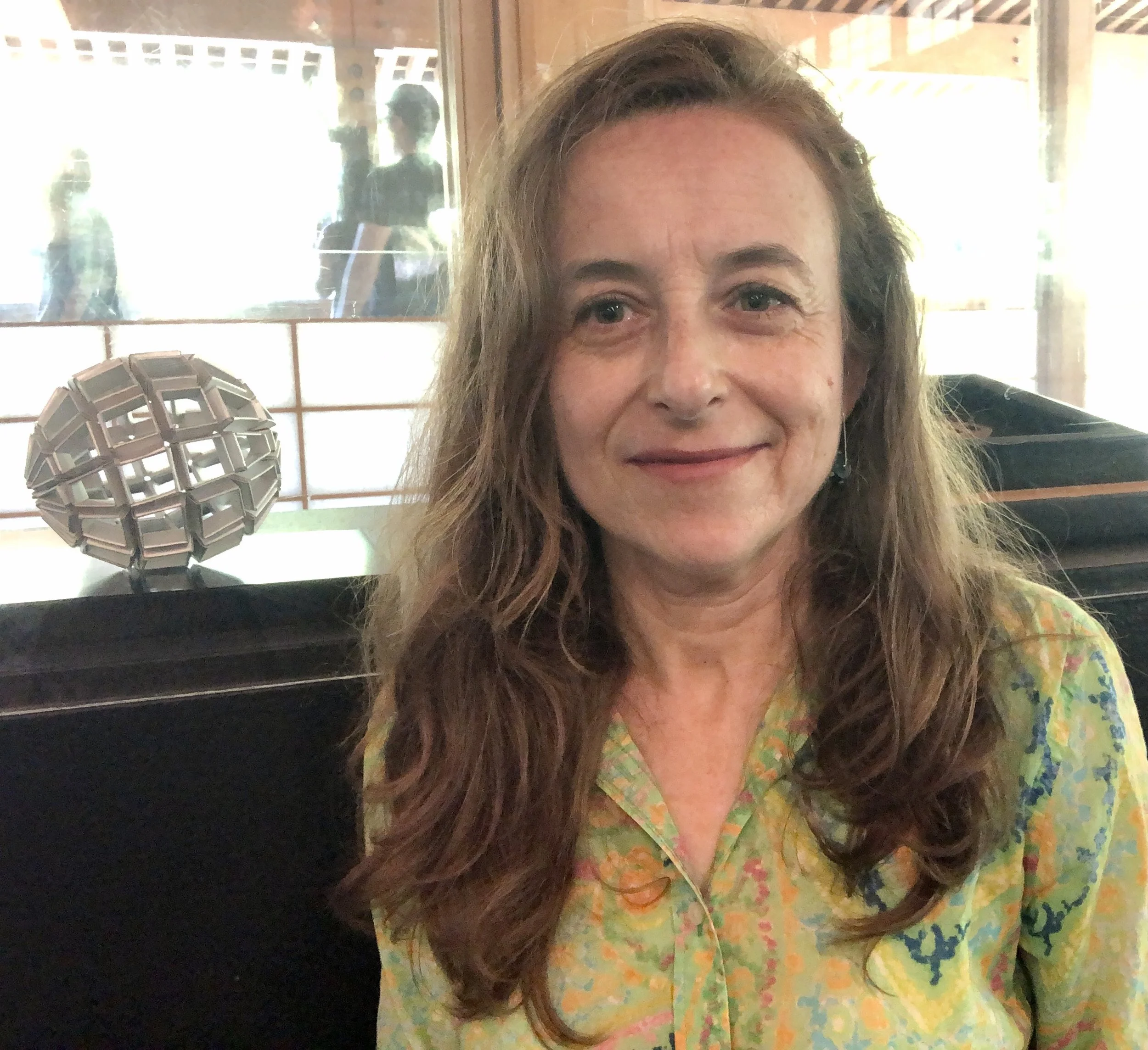 A woman with long, wavy hair sitting indoors, smiling, with a decorative object on the table behind her and a window in the background.