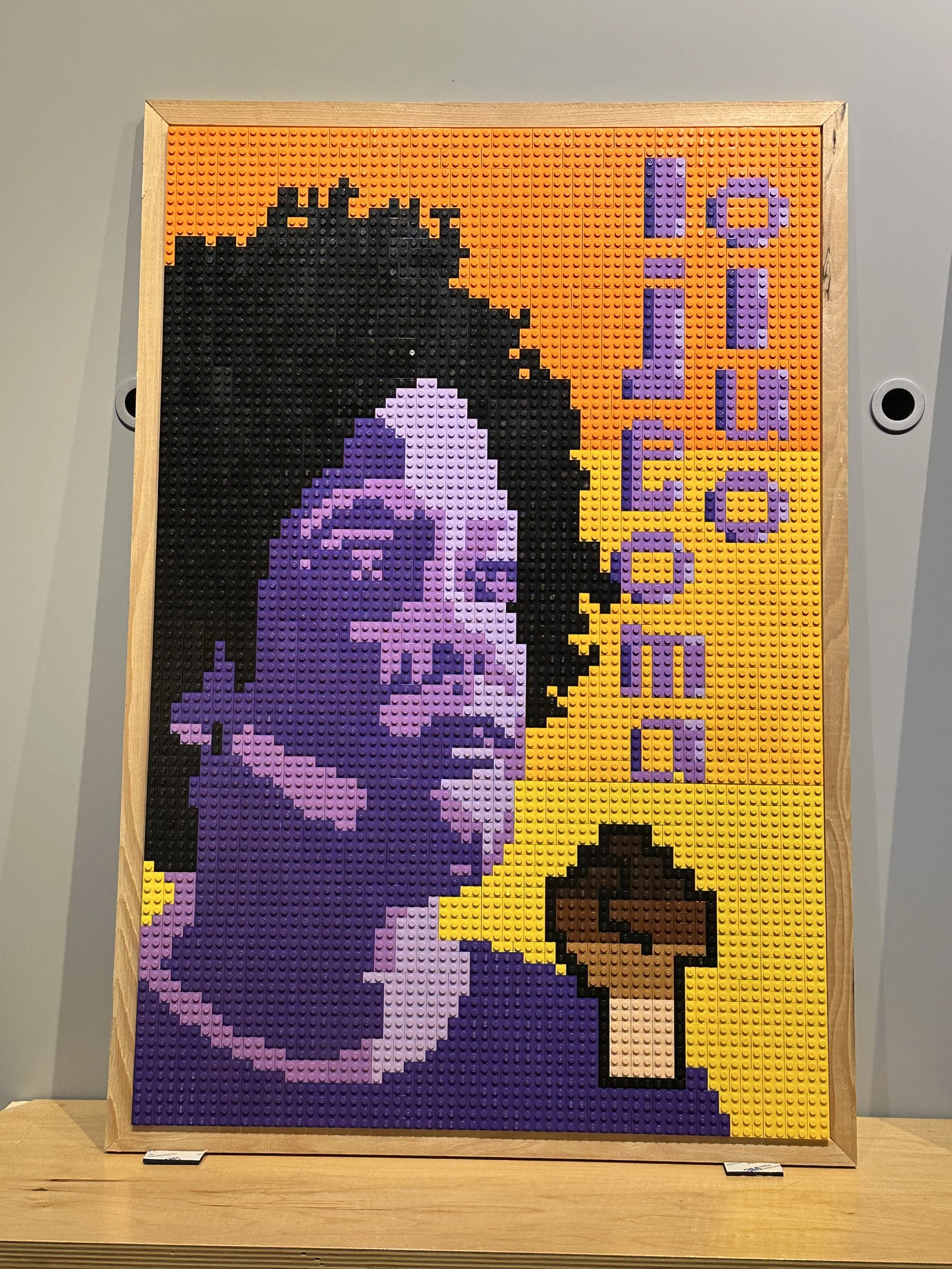 Lego mosaic of a woman with short curly hair, holding a coffee cup, with the text 'Lounge' written vertically in purple on a yellow and orange background.