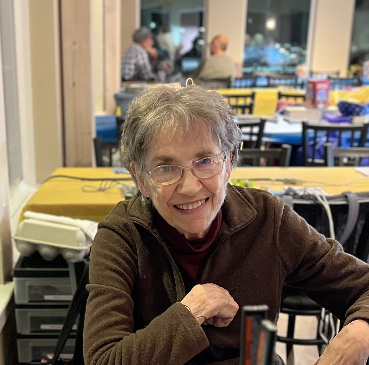An elderly woman with gray hair and glasses smiling at the camera, sitting at a table in a busy indoor setting with people in the background.