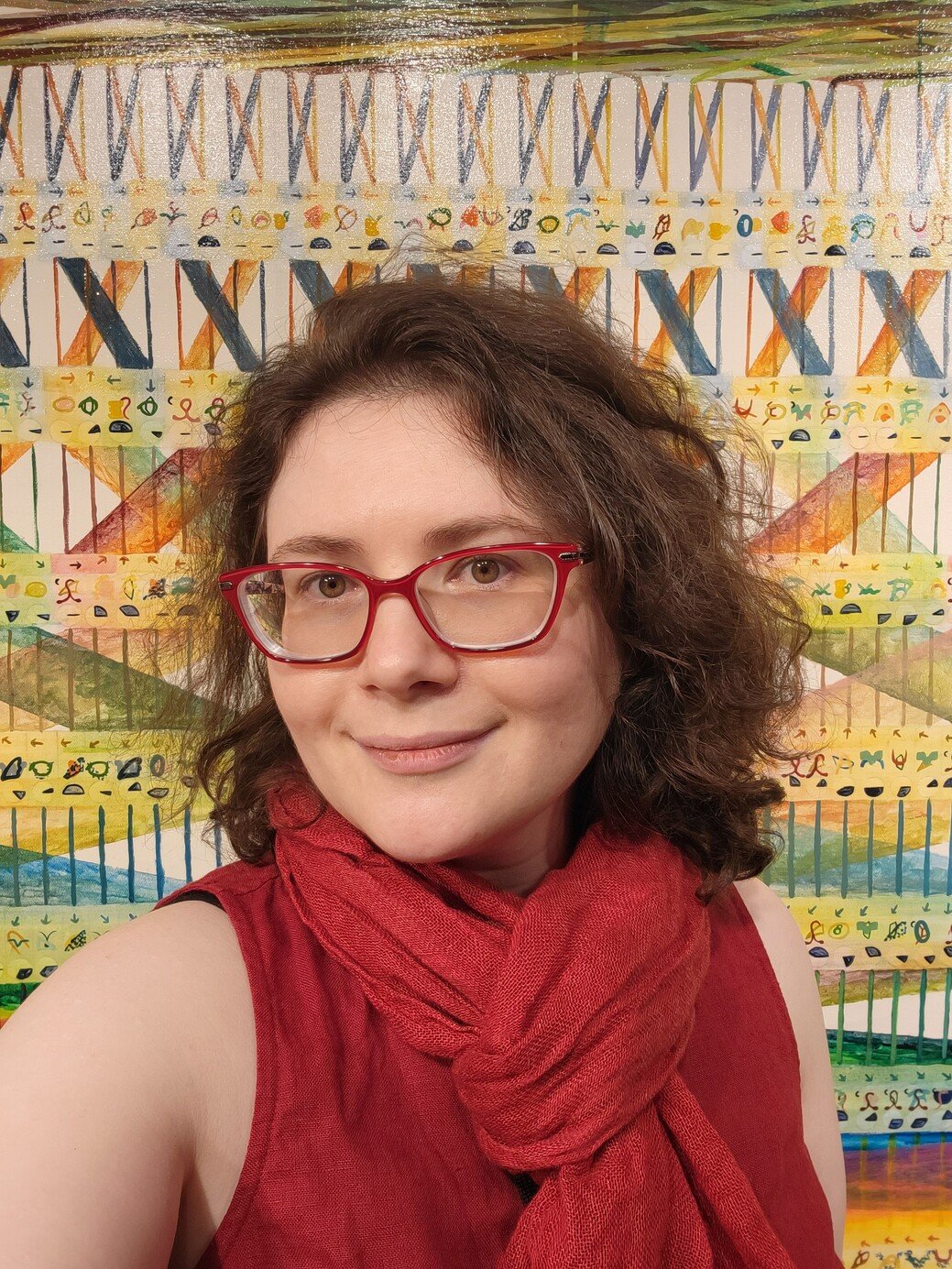 A woman with short curly brown hair, red glasses, and a red scarf and sleeveless top posing in front of a colorful abstract painting.