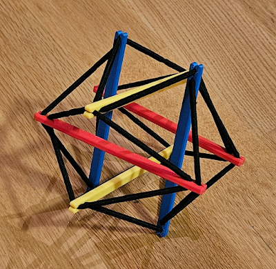 Colorful geometric tensegrity structure made of plastic sticks and black hair ties, resting on a wooden surface.