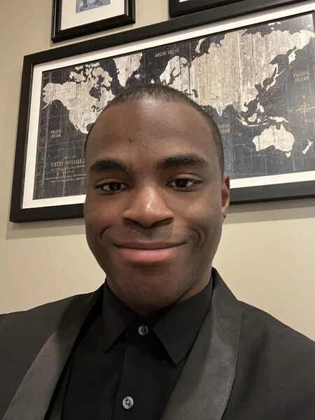 A young person, wearing a black suit and shirt, with short black hair, smiling in front of framed world maps on a wall.