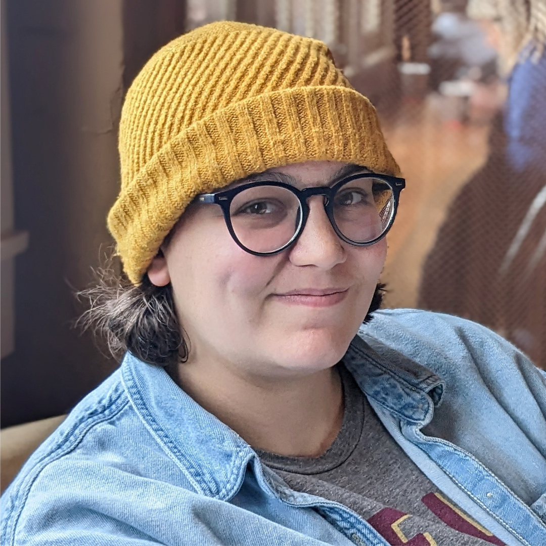 A woman with short dark hair wearing a yellow knit beanie, black glasses, a denim jacket, and a gray t-shirt with red letters, sitting indoors with a blurred background.
