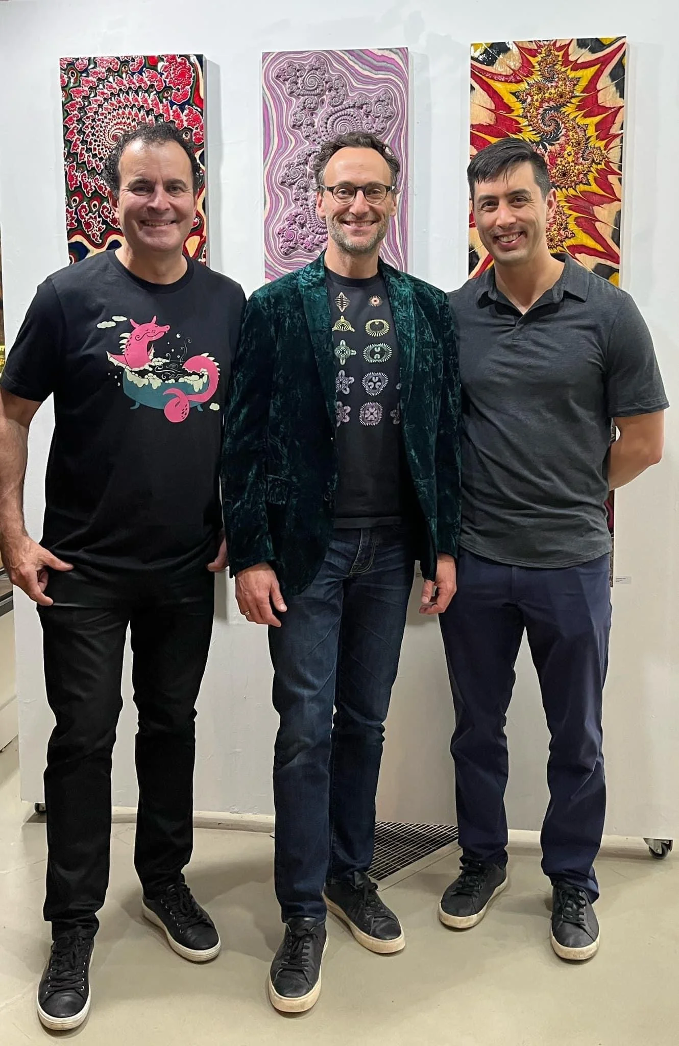 Three men standing together in front of colorful abstract paintings. All three are smiling and dressed casually, with the man in the middle wearing a velvet blazer and glasses.