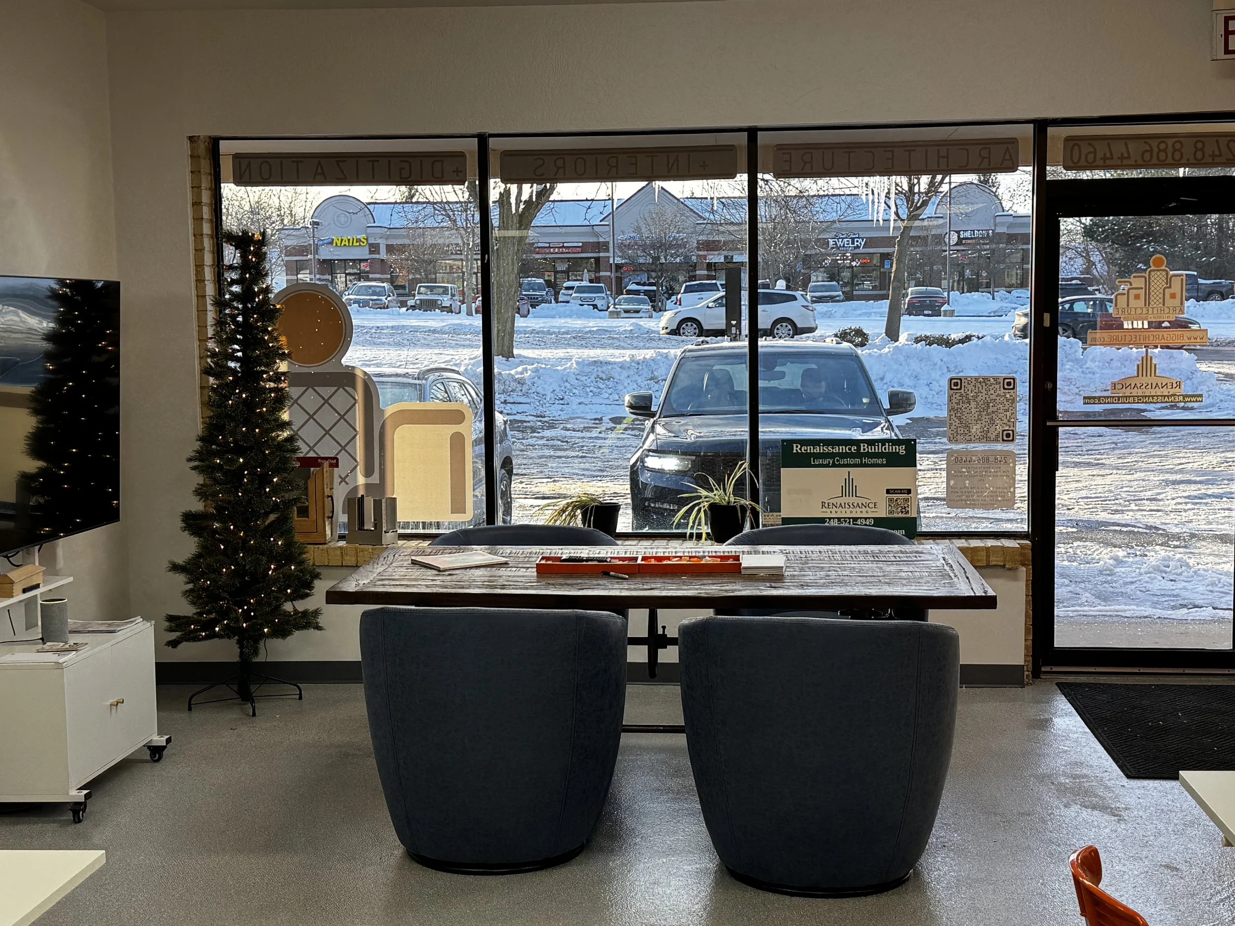Interior of a commercial space with a wooden table, two dark blue chairs, and a decorated Christmas tree. Outside through large windows, snow covers the parking lot and street with cars and retail stores in the background.