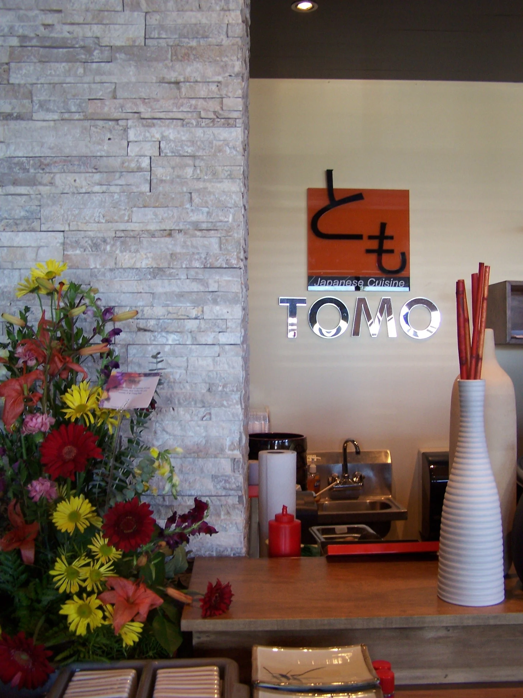 Interior of a Japanese restaurant named TOMO, with a flower arrangement on a counter, a large white vase with red chopsticks, and a kitchen sink in the background. A sign on the wall displays Japanese cuisine.