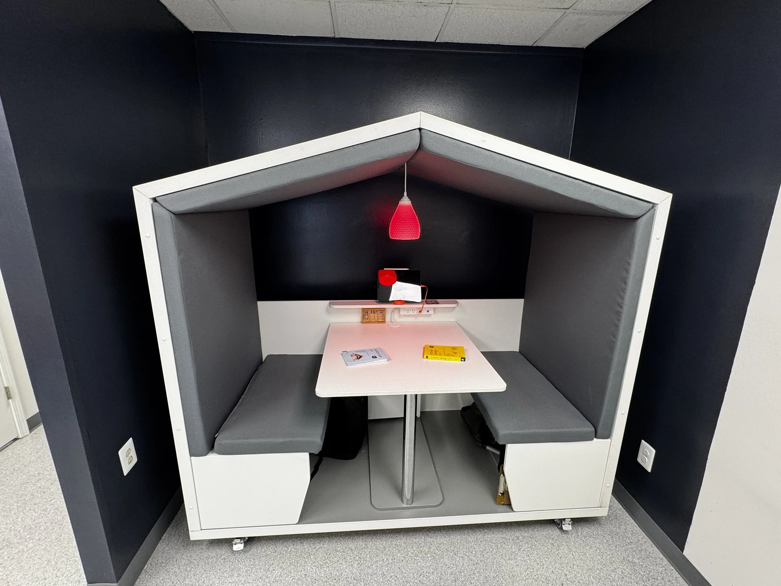 A modern, enclosed study pod with black walls and grey cushioned bench seats on either side of a white table, with a red pendant light hanging above. There are some electronic devices and books on the table, against the wall.