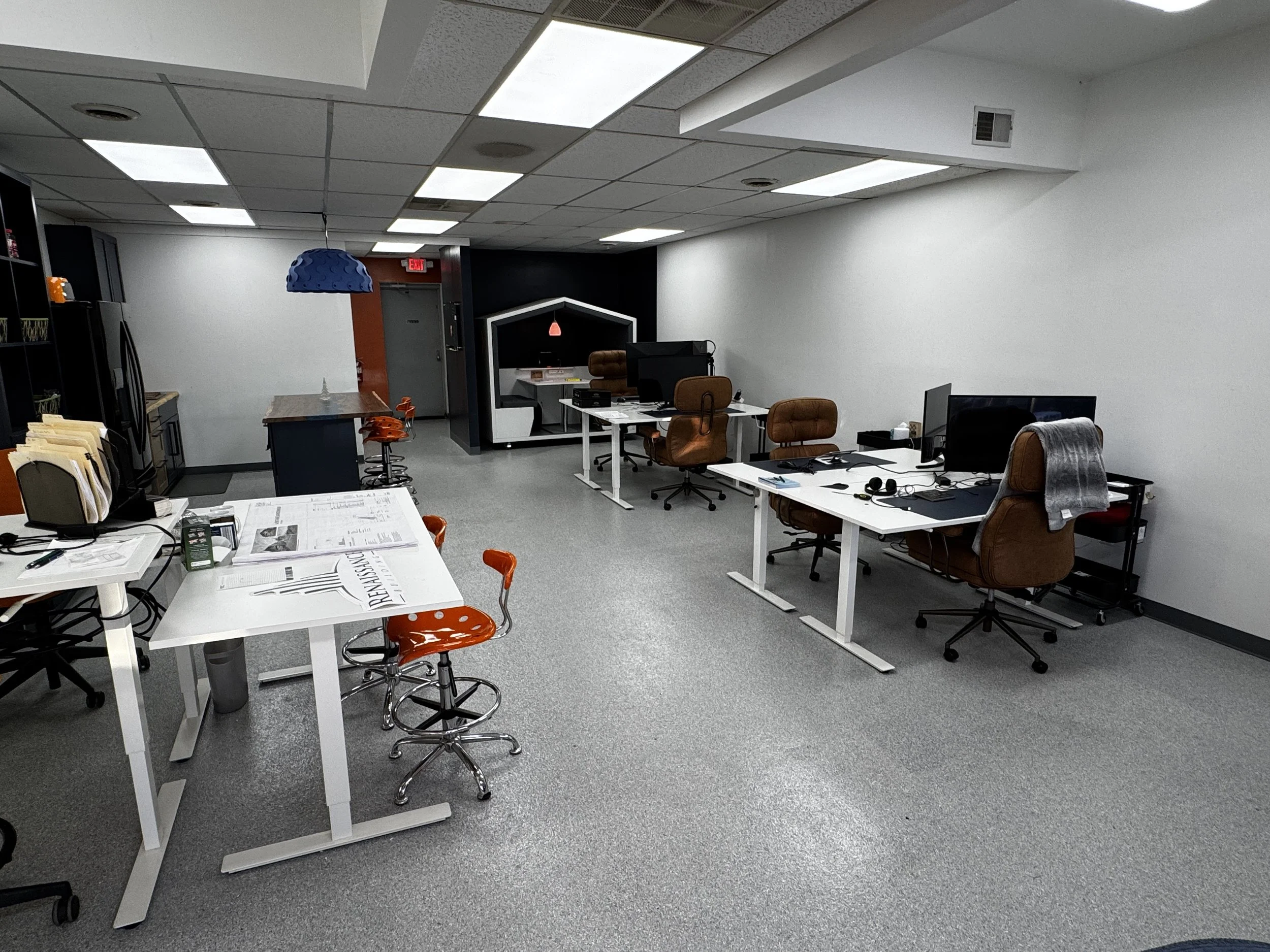 An office space with a row of desks and chairs, some with computer monitors and headphones. There is a small seating area with a high-back chair in a cubicle, a kitchen area with orange stools, and a closed door in the background.