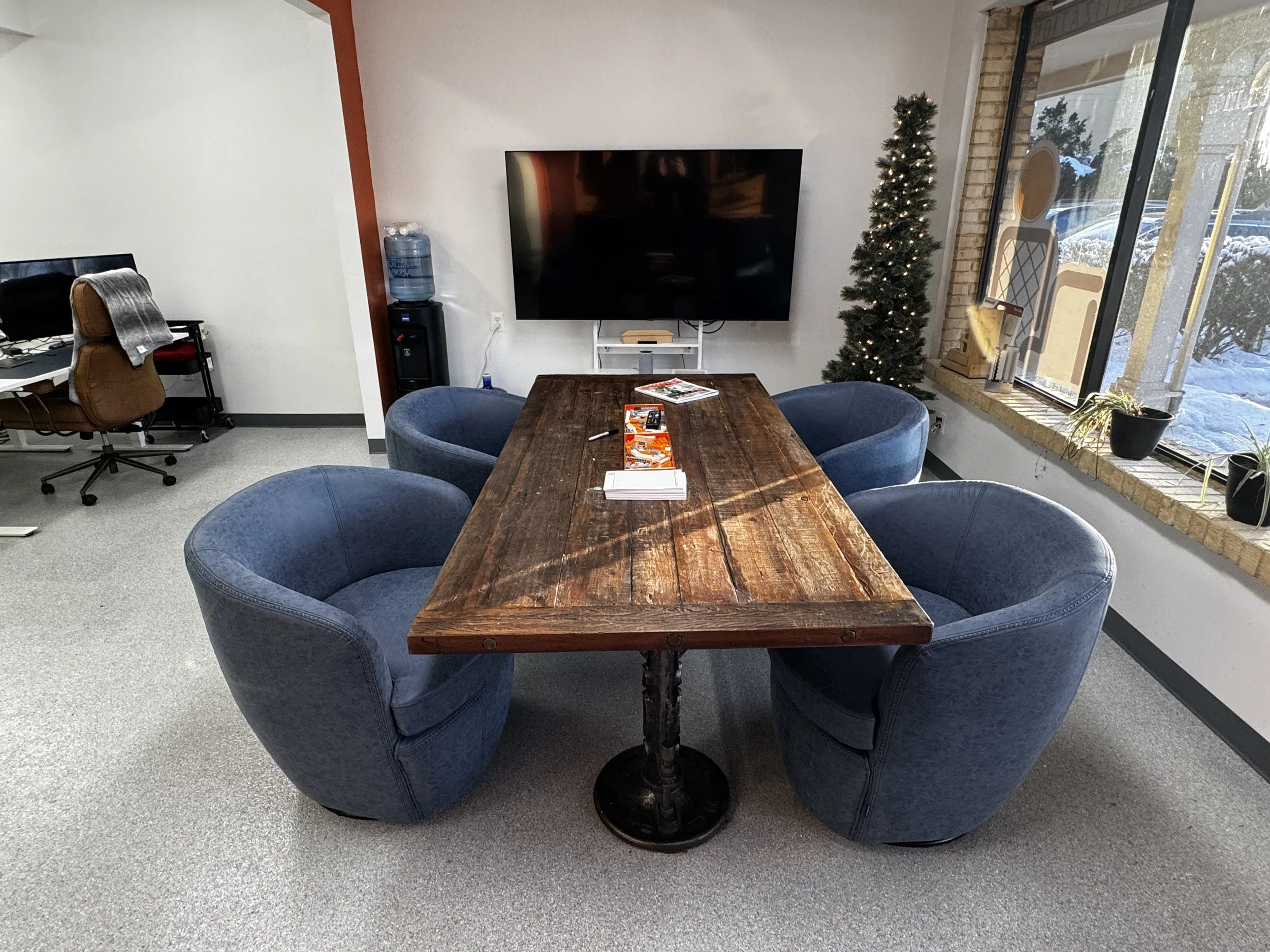 Conference room with a rustic wooden table, four blue upholstered chairs, a wall-mounted flat-screen TV, a Christmas tree, and large windows with sunlight.