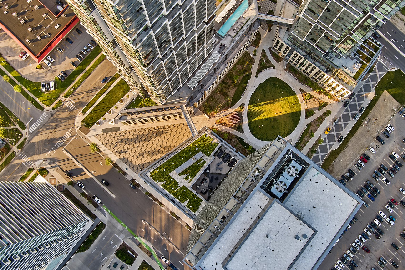 A bird's-eye view of a modern urban area with tall buildings, green spaces, walkways, and parking lots, taken during daylight.