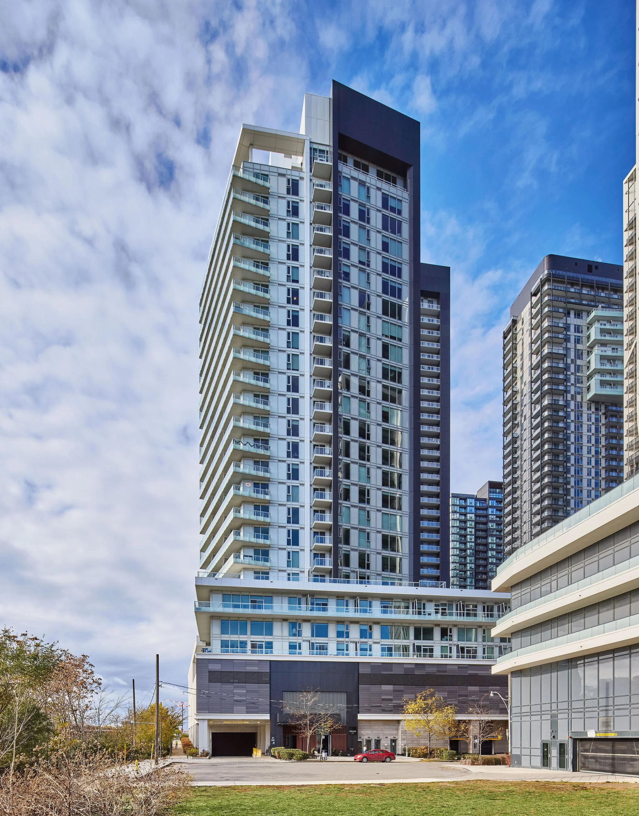 Modern high-rise apartment building with glass windows and balconies, surrounded by other tall buildings under a partly cloudy sky.