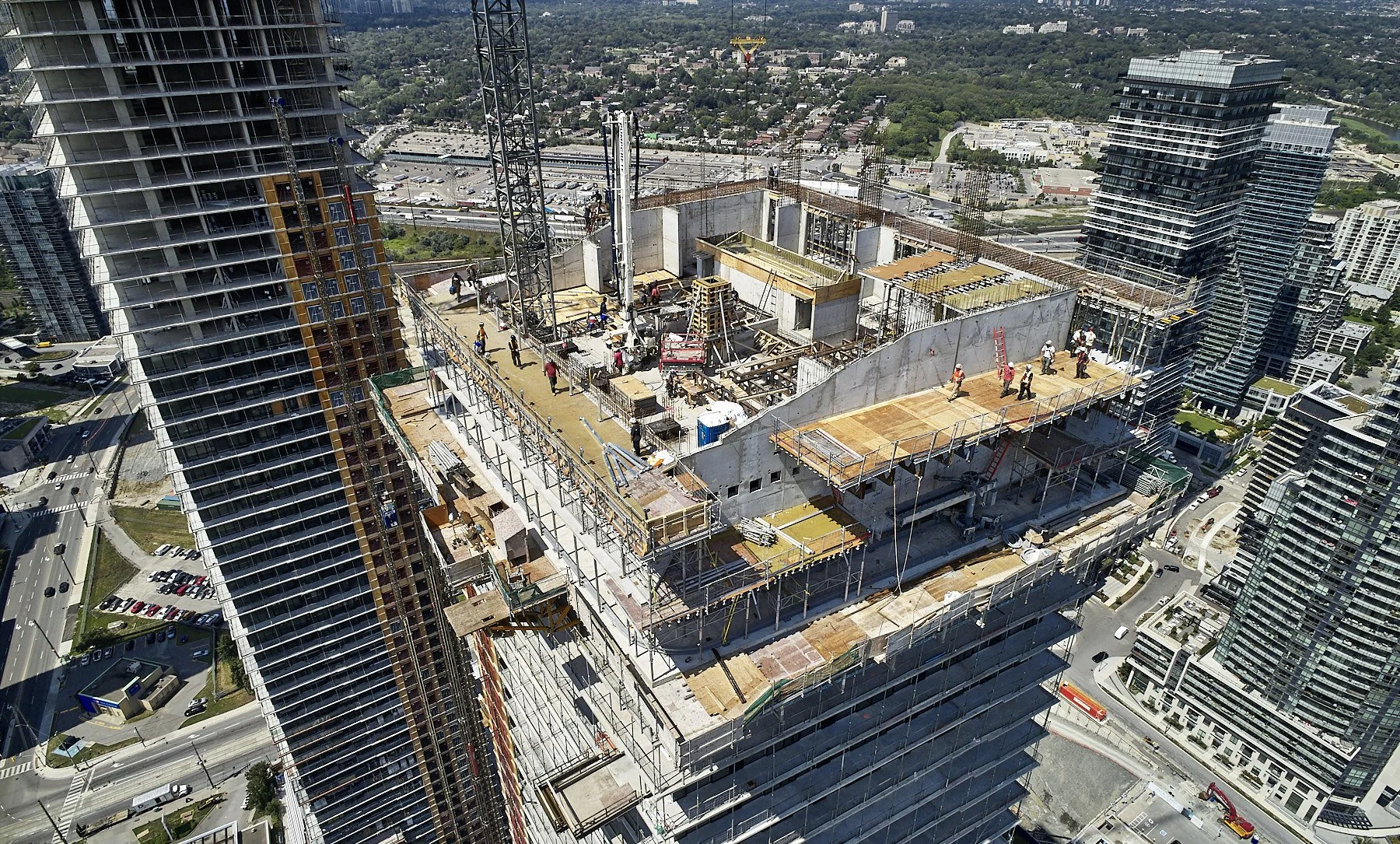 An aerial view of a high-rise building under construction in an urban area with multiple skyscrapers around. Construction workers and equipment are visible on the top floors, with cranes and scaffolding present.