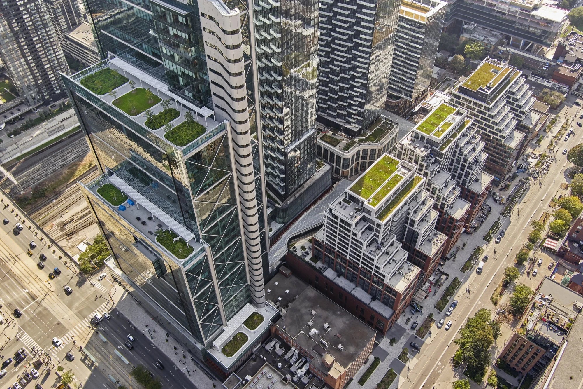 An aerial view of a city block with tall modern glass buildings, some with rooftop gardens, and streets with cars and trees.