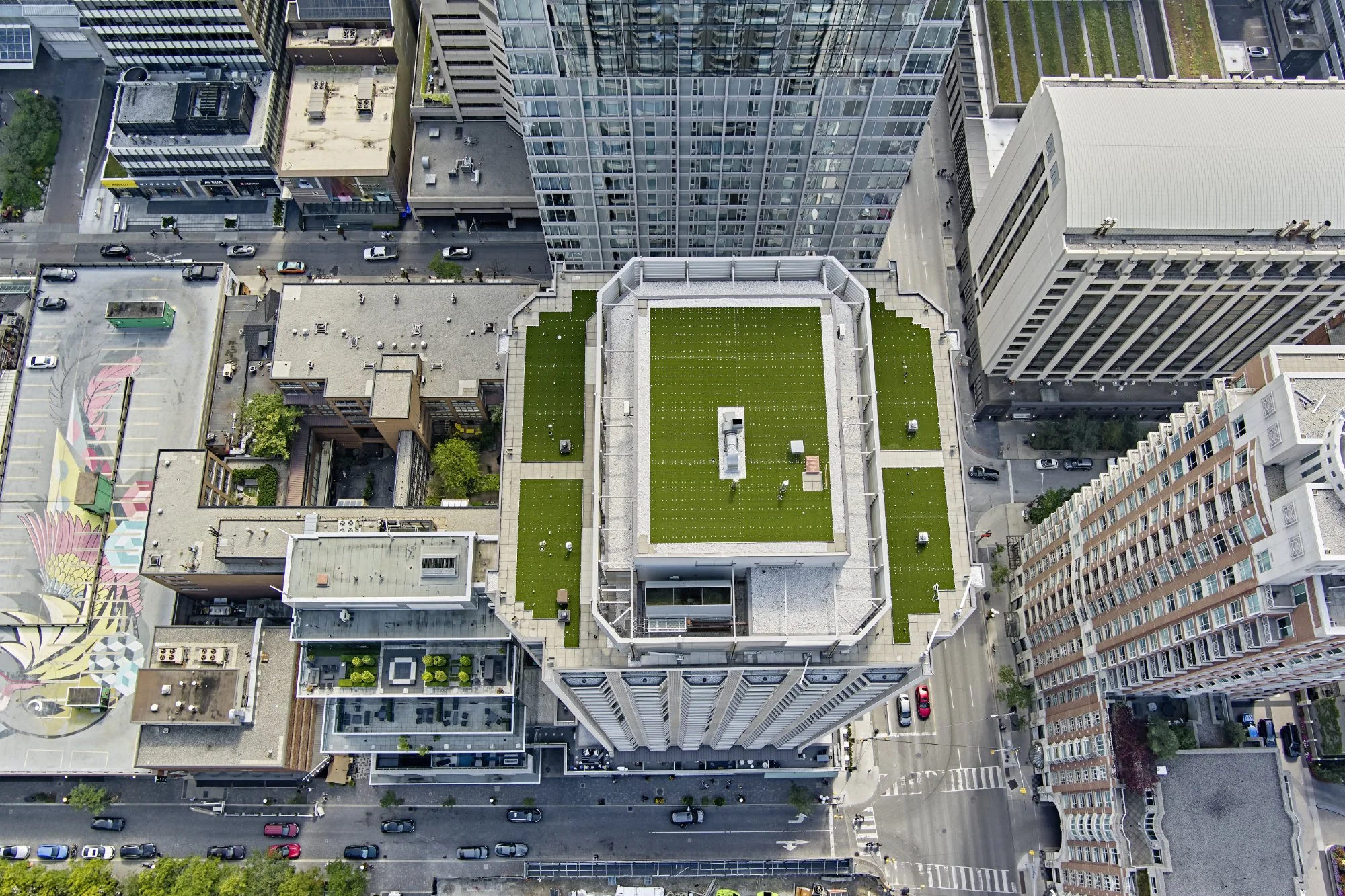 An aerial view of a city skyscraper with a green rooftop, surrounded by neighboring buildings and streets below.