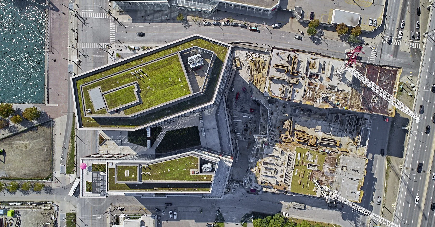 An aerial view of a cityscape with a modern, multi-story building that has green rooftops, adjacent to a construction site with cranes, streets with cars, and a body of water on the left side.