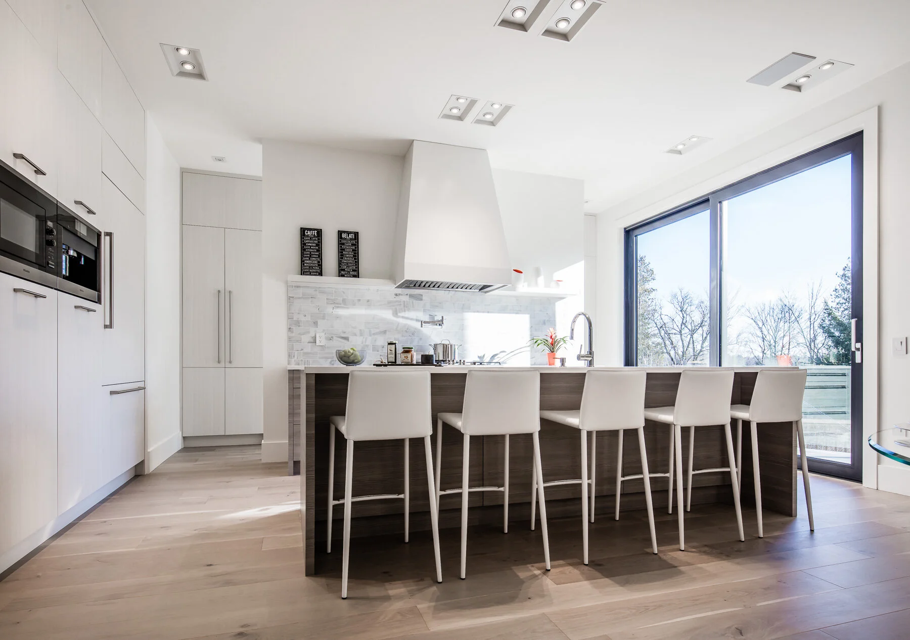 Modern kitchen with white cabinetry, a breakfast bar with beige chairs, a large window facing outdoors, a white range hood, and stainless steel appliances.