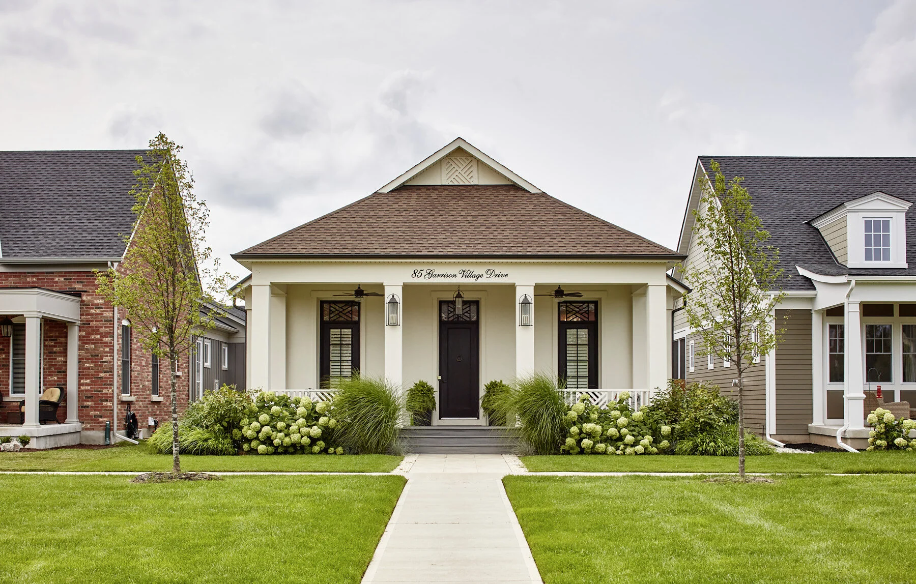 Front view of a house with a white exterior, black door, and a front porch with plants and flowers, flanked by two trees, neighboring houses on either side, with a manicured lawn and sidewalk leading to the entrance.
