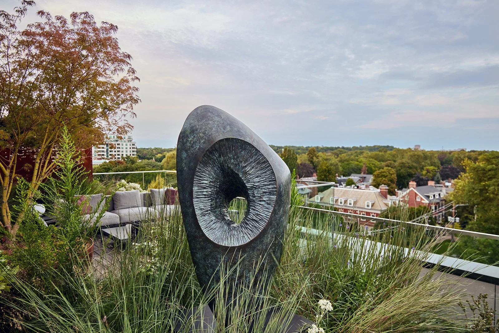 A rooftop garden featuring a large abstract stone sculpture resembling a spiral or circular design, surrounded by green plants, with houses and a tree-lined horizon in the background under a cloudy sky.