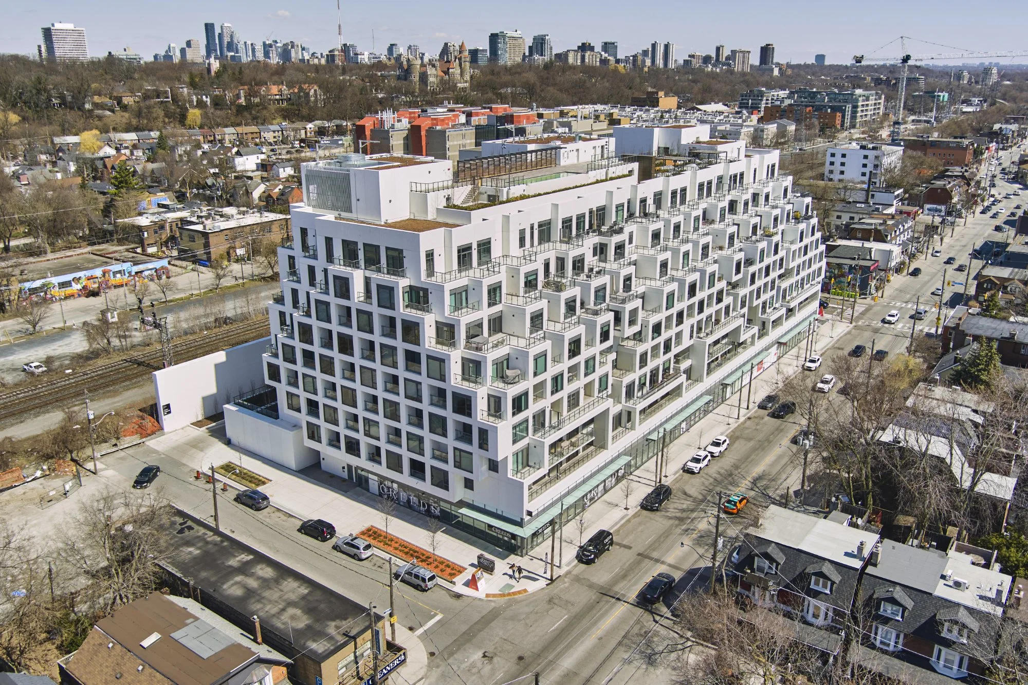 A modern white multi-story apartment building on a city street with cars parked along the curbs, nearby residential houses, and a city skyline in the background.