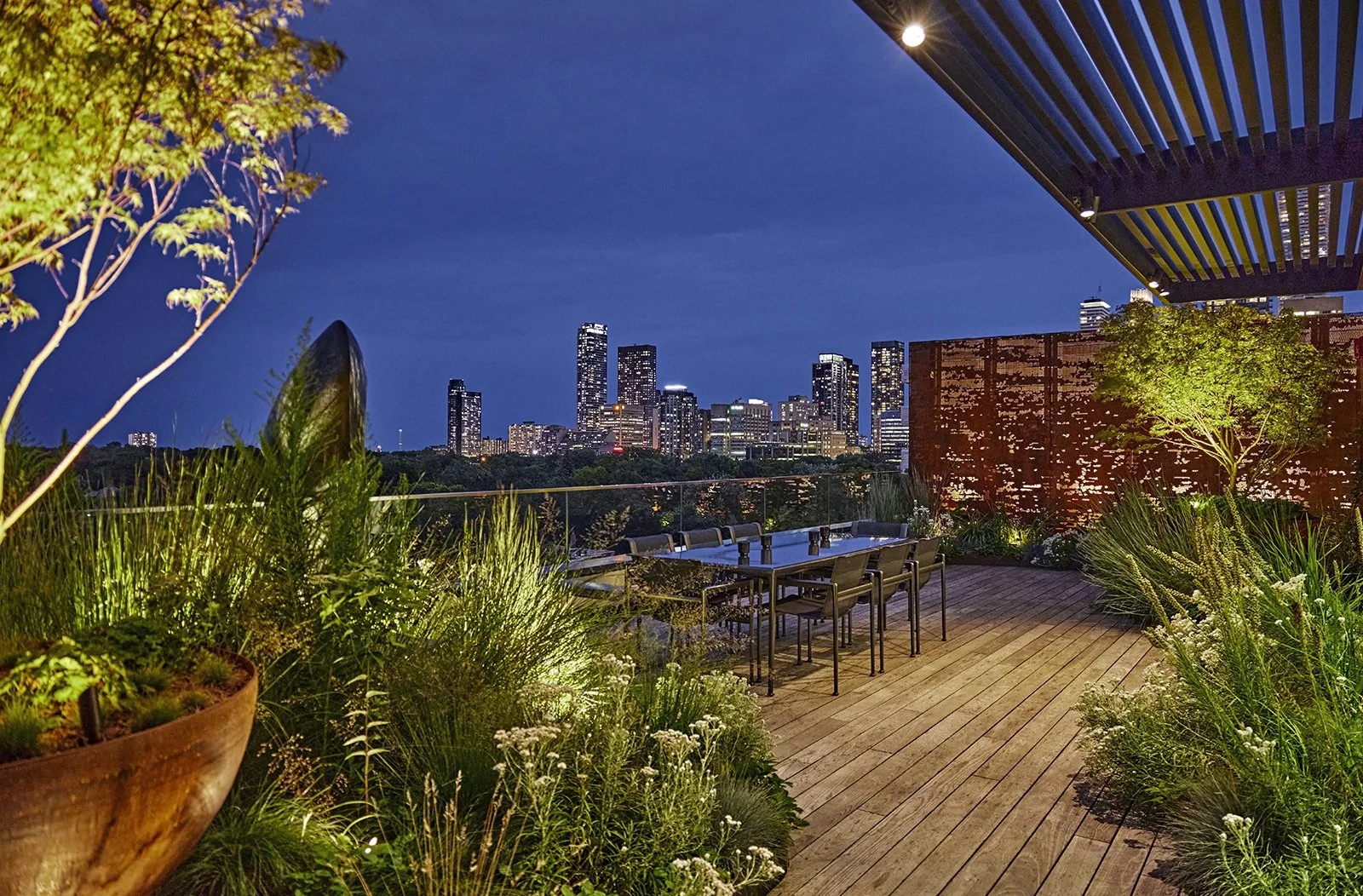 Nighttime city skyline view from an outdoor terrace with wooden flooring, patio table and chairs, greenery, and decorative lighting.