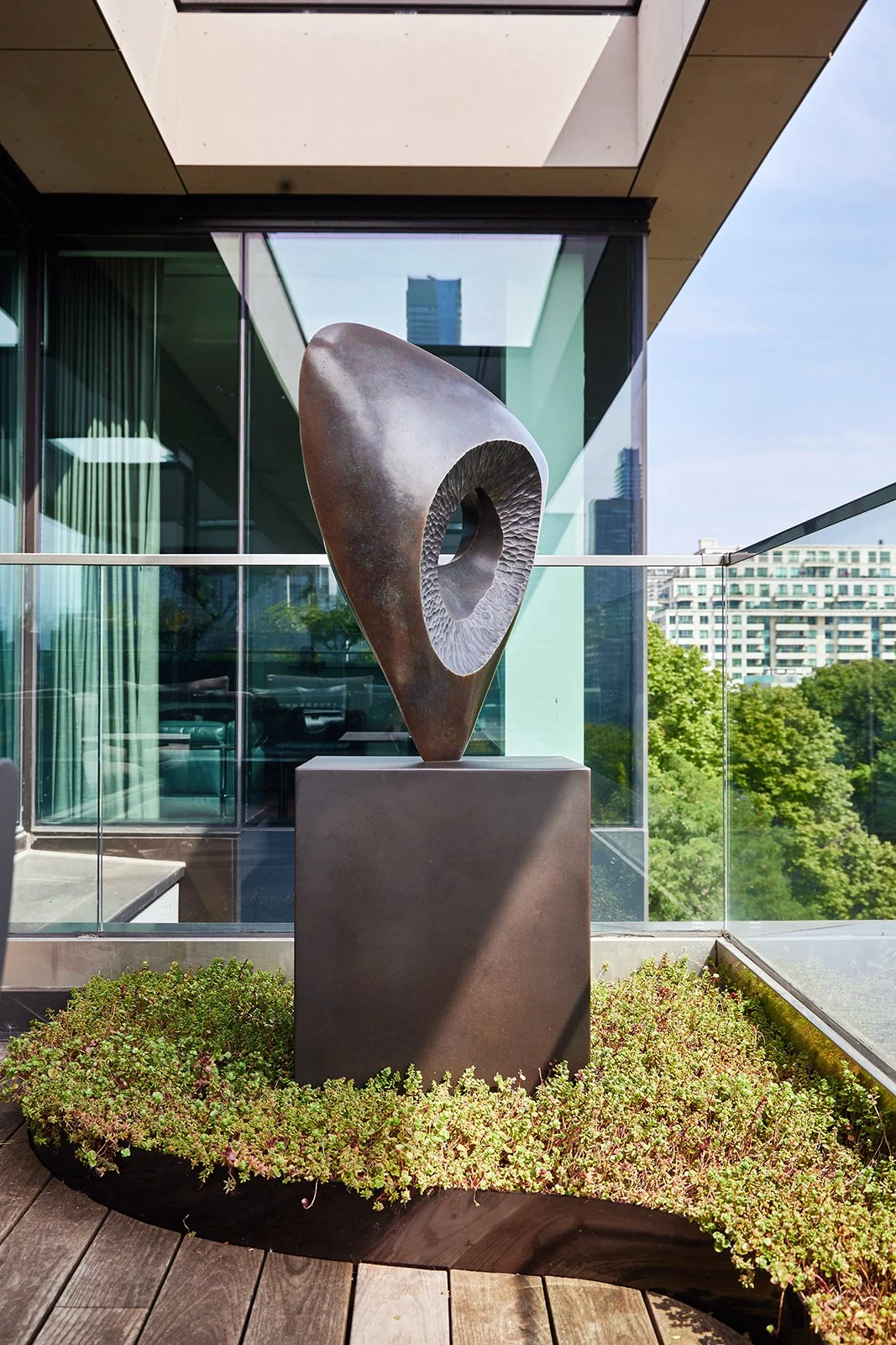 Modern outdoor sculpture of a hollow, abstract, curved form with textured interior, placed on a square pedestal on a balcony with glass railing, greenery, and cityscape in the background.